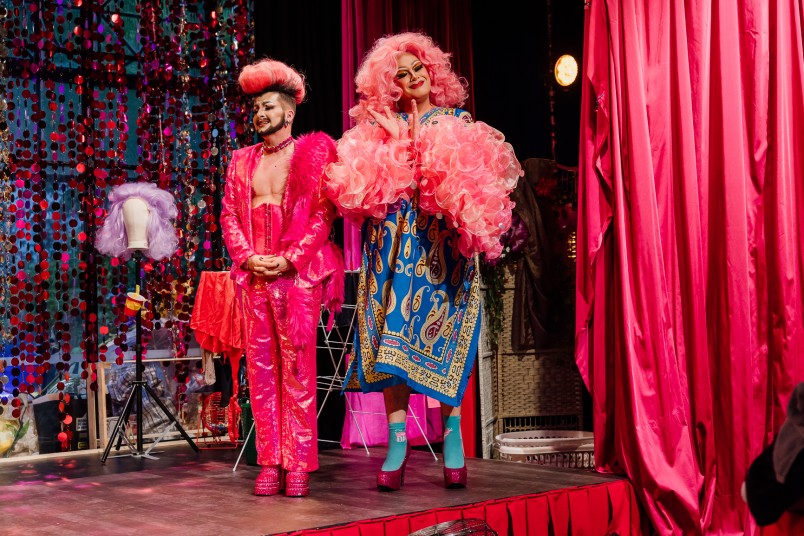 Two of the D.R.A.G performers dressed in full DRAG mid performance, one wearing a pink fringe jumpsuit and the other in a pink wig and feather boa.,