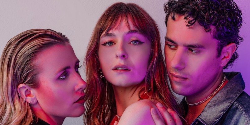 three actors stand together in a clump. the middle actor looks out at the camera, the other two look across her at one another