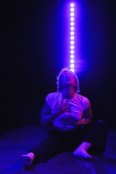An image of an actor on the floor, head back, purple light displaying vertically behind