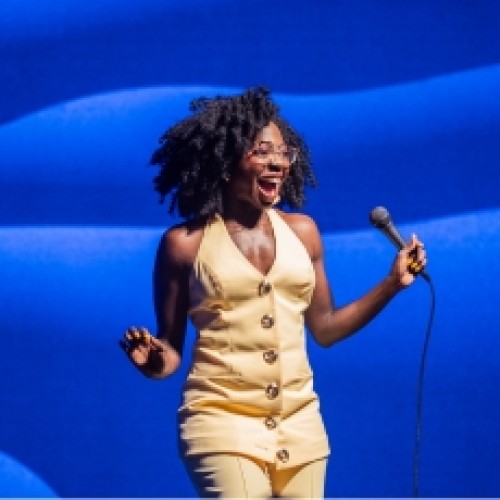 Comedian Anisa Nandaula in a yellow jumpsuit, holding a microphone and smiling with a blue background