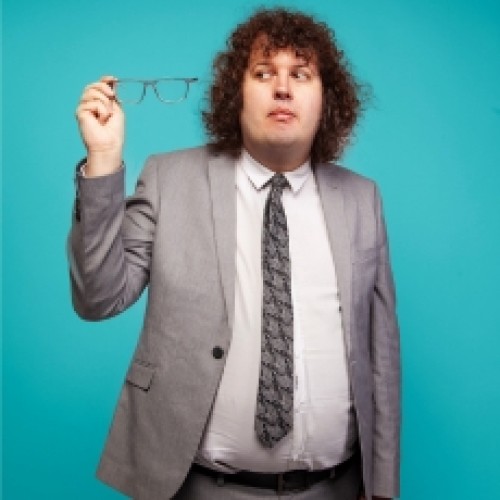 Comedian Ray O'Leary holding his glasses off of his face and peering at them suspiciously, wearing a suit with a bright turqouise backdrop