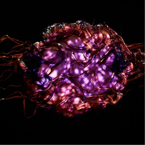 Three performers wearing white contact lenses bathe in purple, shadowy light, nested in what looks like the brown roots of a tree