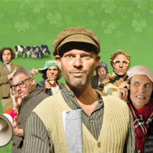 Two actors dressed up as many different characters from a film set, standing in from of a green four leaf clover background with cows behind them. 