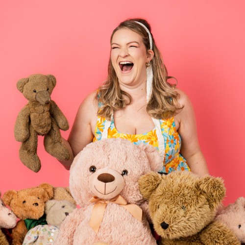 Samantha Hannah holds a teddy bear surrounded by other teddy bears agaisnt a pink backdrop. she is laughing and wears a floral dress and has a ribbon in her blond hair