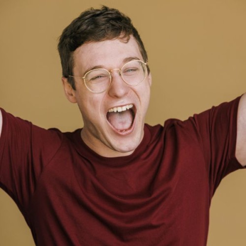 A photo of Anthony Crum, mouth open. He wears a maroon tshirt and gold glasses