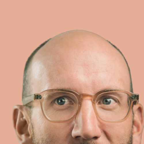 A very close up photo of just Tony's eyes and forehead. He wears glasses and stands agaisnt a light pink background