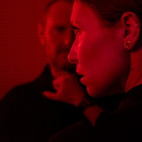 Duotone red image of the side of a woman's face, she is looking to the left. Beyond her is a man facing the viewer, his face is blurry becasue the focus is on the woman.