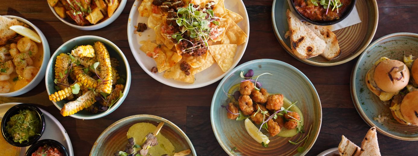 Our Food Menu - A variety of plated dishes on a wooden table, including nachos with meat topping, grilled corn, shrimp, prawn, sliders, and dipping sauces, arranged for a shared dining experience."