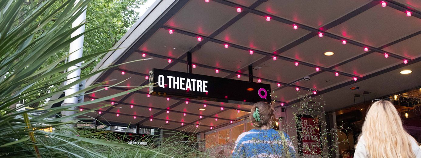 The image shows the entrance to Q Theatre, a performing arts venue. The sign "Q THEATRE" is illuminated under a canopy with a grid of pink lights. Two people are seen walking toward the entrance, one wearing a blue sweater with a hair clip and the other with long blonde hair. Green plants frame the scene, and trees in the background suggest an urban outdoor setting.