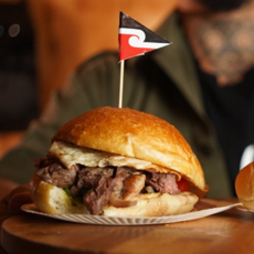 An image of a very meaty burger with a Maori Flag stuck into it.
