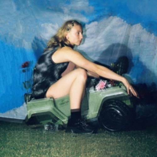 A women with hair clipped back, wearing a black tie dye play suit and black shoes, looks off pensively whilst sitting on a toy army grey truck on fake grass in front of a background that looks a bit like sky / mountains.