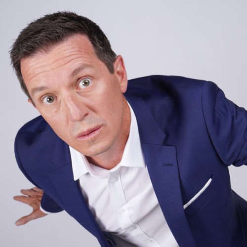 Photo of Rove McManus. He wears a blue suit and looks up into the camera.