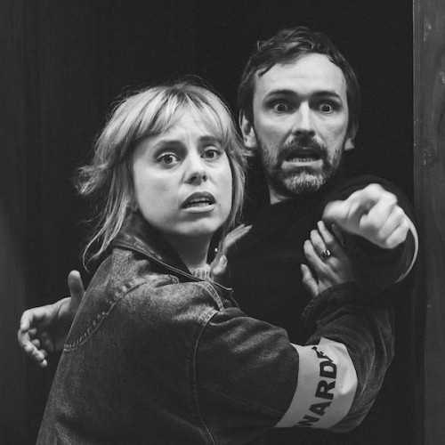A greyscale photo of two people looking frightened. One points at someting behind us. On the wall to the right we notice the shadow of a giant werewolf...